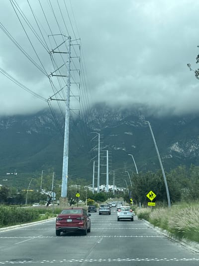 The image displays a multi-lane asphalt road in Ciudad Santa Catarina, Mexico, oriented towards a mountainous backdrop partially obscured by low-hanging clouds. The road surface features a grid pattern of white reflective studs. On the left, several tall metallic utility poles carrying numerous power lines extend into the distance. Modern streetlights with curved poles are visible on both sides of the road. Two yellow, diamond-shaped warning signs, each depicting two pedestrians and the word "PRECAUCION" (caution), are present on the right and left verges, indicating school crossing zones. The roadside verges are lined with sparse green vegetation and shrubs. In the foreground, a dark red sedan is positioned in the left lane, moving away from the viewer. Further down the road, a silver/white hatchback is visible in the right lane, also moving away. Additional vehicles are discernible in the distance, indicating moderate traffic flow. White buildings are present at the base of the mountains. No individuals are visible.