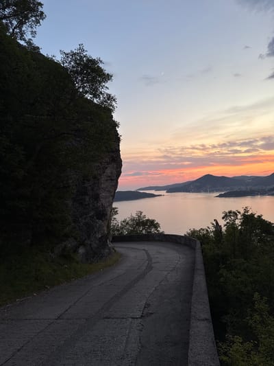 The image depicts a scenic sunset view over a body of water, likely the Adriatic Sea, from a high vantage point. In the foreground, a winding asphalt road curves to the right, bordered by a low concrete barrier on the right and a steep, rocky cliff face covered in dark green foliage on the left. The road shows signs of wear, with visible cracks and tire marks. Further in the distance, mountains form a silhouette against the vibrant, colorful sky, characterized by hues of orange, pink, and pale blue, with a faint crescent moon visible. The water reflects the warm colors of the sunset. Several trees with dark green leaves are scattered throughout the scene, some emerging from the cliff face and others on the slopes leading down towards the water. A distant, faint structure or building complex is visible on one of the mountainous peninsulas jutting into the sea. No people are clearly identifiable in the image. The overall atmosphere is serene and picturesque, characteristic of a twilight view from a coastal mountain road. The location is identified as Podličak, Montenegro.