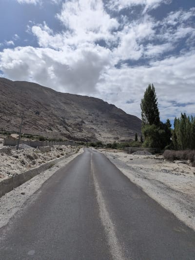 The image depicts a road in Sasoma, India, stretching towards a mountainous landscape under a partially cloudy sky. The road is paved and asphalt, with a white line dividing the lanes. On either side of the road, there is dry, sandy terrain with some scattered rocks and vegetation. A concrete drainage channel runs along the left side of the road. In the distance, a rugged, barren mountain dominates the background. A cluster of green trees is visible to the right of the road, contrasting with the arid surroundings. There are no people or vehicles visible in the image. The overall scene suggests a remote, dry environment with a road traversing through it.
