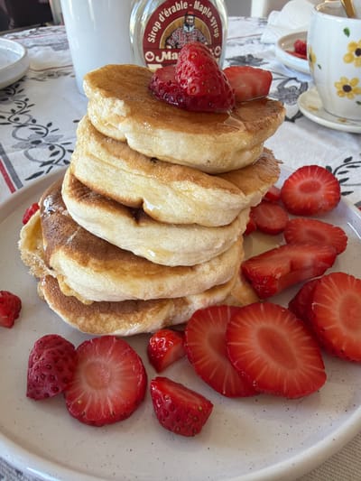 This image captures a prepared breakfast or brunch scene, situated in Rueil-Malmaison, France. The primary subject is a tall stack of approximately seven fluffy, golden-brown pancakes, heavily drizzled with clear, amber maple syrup that glistens and drips down their sides. A single whole, ripe red strawberry rests atop the stack, accompanied by a partial strawberry slice.
The pancakes are presented on a round, light-colored ceramic plate with a subtle speckled texture. Scattered around the base of the pancake stack on the plate are numerous additional fresh red strawberries, varying in presentation from whole berries to halves and multiple slices, all displaying vibrant red flesh and small, visible seeds.
In the background, a glass bottle of maple syrup is partially visible to the upper right. Its label clearly reads "Sirop d'érable - Maple syrup" and features a graphic of a man with a beard and a hat, wearing a plaid shirt. Adjacent to the syrup bottle, a white ceramic teacup or coffee mug adorned with a distinctive yellow floral pattern (likely sunflowers) and its matching saucer are partially visible. To the left background, a plain white ceramic bowl is also partially visible. All items are arranged on a white tablecloth patterned with repeating dark grey or black stylized floral and foliate motifs. The lighting is bright and even, indicating an indoor setting. No individuals or actions are depicted; the scene shows a static display of food.