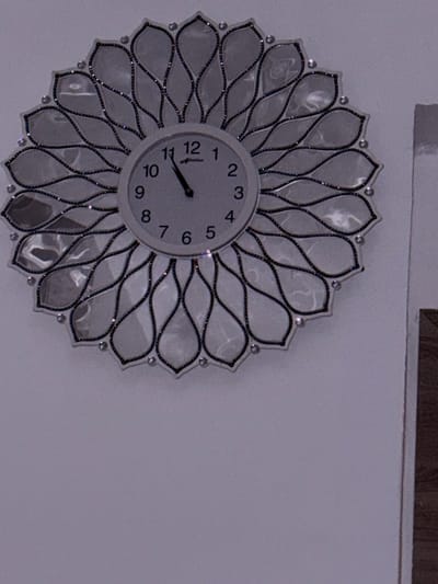 A wall clock with a decorative, floral-like frame is mounted on a light-colored wall. The clock face is round and white, with black Arabic numerals and black hour and minute hands. The hour hand points slightly past the 11, and the minute hand points to the 5. The frame of the clock is made of mirrored, petal-shaped elements outlined with black and adorned with small, clear rhinestones. The clock is centrally located on the wall, with a portion of a dark wooden surface visible to the right of the clock, possibly a door frame or furniture. The lighting suggests an indoor setting. The image does not provide information about the city of Evere, Belgium.