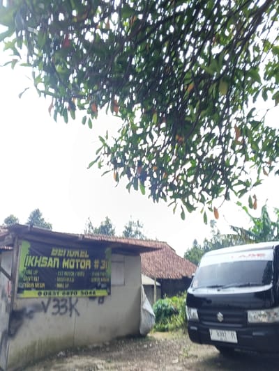 A small automotive repair shop, "Bengkel Ikhsan Motor #31," provides services such as tune-ups, oil changes, engine overhauls, paint jobs, modifications, and spare parts. The shop's contact number is 0831 6870 5048. Graffiti, "V33K," is visible on the shop's wall. A black Suzuki minivan is parked nearby on a dirt road under tree foliage, partially obscuring the shop and a background house with a tiled roof. The overall setting appears to be rural.