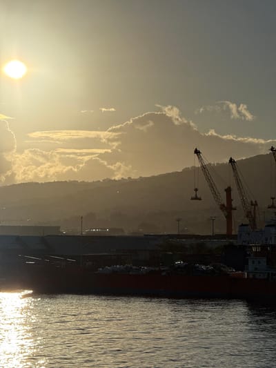 The image depicts a port scene in Papeete, French Polynesia, at either sunrise or sunset. A bright, low sun is visible on the far left, casting a golden light across the sky and creating a strong reflection on the rippled water in the foreground. Hazy, silhouetted mountains form the background, topped by scattered cumulus clouds illuminated by the low sun. In the midground, a dark-hulled cargo ship is moored, its deck laden with a pile of various materials. Behind the ship, several low-rise port buildings and utility poles are visible. To the right, three large industrial cranes stand silhouetted against the sky and mountains, one of which has a container spreader attachment hanging from its arm. No individuals are discernible in the scene.