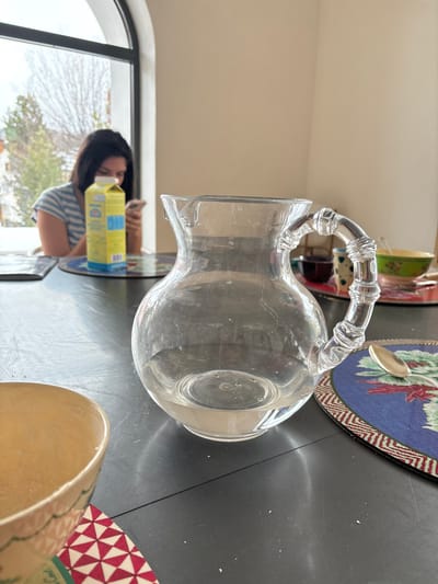 A glass pitcher with a clear liquid and a bamboo handle sits on a gray table in a dining area. It is located next to a colorful placemat and a spoon. A partially visible, empty, yellow bowl with a red and white patterned coaster sits next to the pitcher. A blurry figure of a woman sits in the background. The woman is wearing a blue and white striped shirt and is looking down at a mobile phone.  There is a yellow box to the left of the woman. The table is covered in a grey tablecloth. The background is a white wall with an arched window. The window is open and shows a blurry view of a green landscape. The location is a home in Celerina/Schlarigna, Switzerland. The coordinates are 46.514, 9.859.