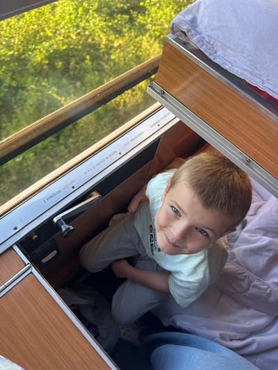 A young boy with blond hair and blue eyes is looking up at the camera, smiling. He is wearing a light blue t-shirt and grey sweatpants. He is sitting inside what appears to be a camper or RV, with wooden interior paneling visible. A window with green foliage visible outside is to his left. A white duvet or blanket is stacked on a shelf above him. The image does not provide enough information to determine the location in Valjevo, Serbia.