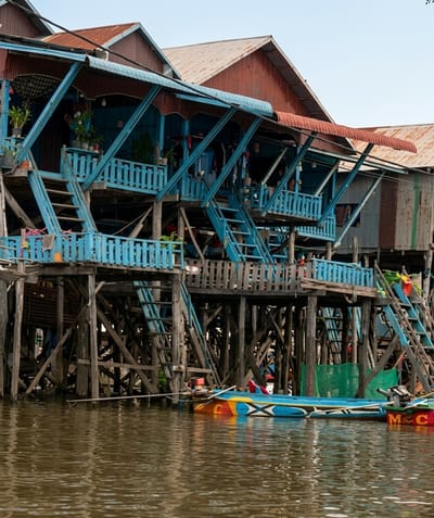 In Soutr Nikom, Cambodia, a stilt village is depicted with weathered wooden structures resting above calm, brown water. The buildings feature brightly painted blue railings and balconies, contrasted with corrugated metal roofs in shades of red and brown. Multiple staircases, some painted blue, ascend to the elevated structures, with laundry visible hanging from some of the balconies. At the base of the buildings, a vibrantly colored boat with blue, yellow, and red sections, and an outboard motor, is moored to the left side of the frame. The overall scene captures the unique architecture and lifestyle of a waterside community.