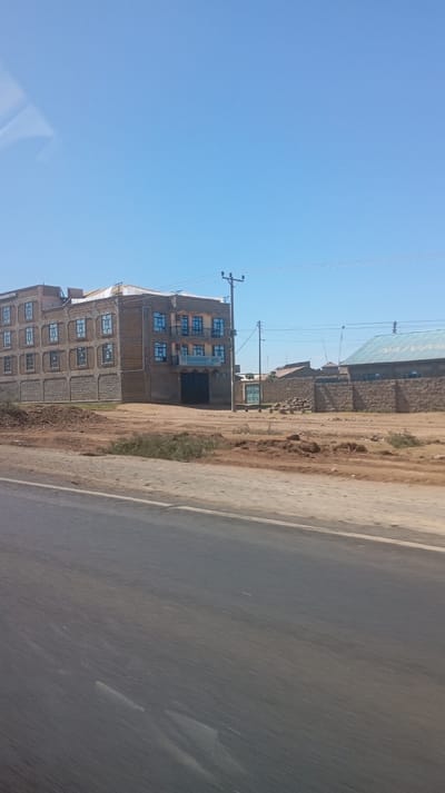 A building, likely a hotel or inn, stands prominently along a road. It's three stories tall with a tan brick facade and blue-framed windows. A sign reading "Summer Hill" is visible. Power lines stretch across the clear blue sky. In the background, a lower building with a corrugated metal roof and stone walls is visible, separated by a dirt lot. The foreground is dominated by the asphalt road, showing a blurred, fast-moving perspective. The lighting indicates a bright, sunny day.