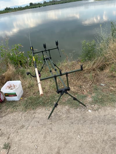 The image depicts a fishing setup by a body of water, likely a lake or reservoir, under an overcast sky. In the foreground, a fishing rod is placed on a tripod rod rest. A white bucket, containing what appears to be bait or chum, sits to the left of the rod rest. The surrounding area is covered in dry grass and dirt. In the background, the calm water reflects the sky and distant trees. The scene appears to be set in a natural environment, possibly on the outskirts of the city of Voyvodino, Bulgaria, given the mention of the location. There are no people visible in the image, and the focus is on the fishing equipment and the natural surroundings.