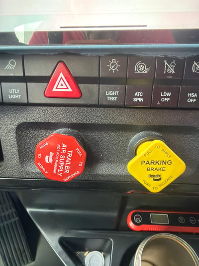 The image displays the interior of a truck's control panel, specifically focusing on the dashboard area. Prominently featured are two large, distinct control knobs: a red octagonal knob labeled "TRAILER AIR SUPPLY NOT FOR PARKING" with "PUSH TO SUPPLY" and "PULL TO EVACUATE" instructions, manufactured by Bendix. To its right is a yellow diamond-shaped knob labeled "PARKING BRAKE" with "PUSH TO RELEASE" and "PULL TO APPLY" instructions, also manufactured by Bendix. Above these knobs, a row of smaller buttons is visible, including a red hazard light button with a white triangle, and other buttons with symbols and text such as "UTLY LIGHT," "LIGHT TEST," "ATC SPIN," "LDW OFF," and "HSA OFF." The background shows a textured, dark surface typical of truck interiors, with a faint reflection of what appears to be a person in red clothing on the upper portion of the dashboard. The image is a close-up shot, capturing the detail of the controls. The setting is not identifiable as being in Lebanon, United States of America.