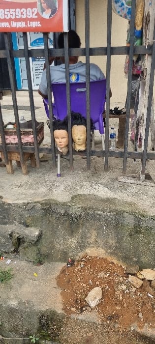 The image presents an outdoor scene in Palmgrove, Ilupeju, Lagos, Nigeria, viewed through a dark vertical metal fence in the foreground. A person, seen from the back, is seated on a purple plastic chair behind the fence. To their left, two light-skinned mannequin heads, each wearing a dark wig, are displayed on individual stands, visible through the fence. Adjacent to the left mannequin head, a small wooden stool supports a brown wicker basket. A plastic water bottle rests on the ground to the right of the seated person. Above the scene, a partially visible red and white sign displays the number "8559392" and the text "Palmgrove, Ilupeju, Lagos." The immediate foreground consists of a cracked concrete surface with a significant mound of reddish-brown dirt, rocks, and miscellaneous debris. In the background, a light-colored building with a dark entryway is visible.