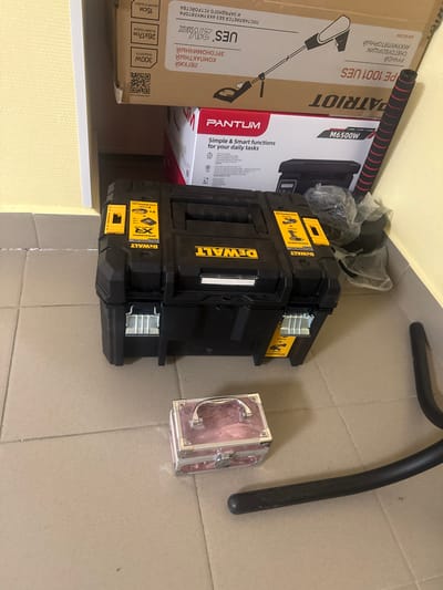 The image depicts a corner of an indoor space, likely a storage area or room, with light beige textured walls and a light beige tiled floor. The scene is cluttered with several items, suggesting a somewhat disorganized environment.

In the center of the frame, a black DeWalt tool case is prominently displayed. It is a rectangular, hard-sided case with yellow accents and branding. The case appears to be closed. To the right of the tool case, a black metal object, possibly part of a rack or stand, curves into the foreground.

Behind the tool case, two cardboard boxes are stacked. The top box is light brown and appears to be a product box for a "PATRIOT" branded item, with Cyrillic text and an illustration of a cleaning device. Below it, a white and red box with "PANTUM" branding is visible, likely containing an appliance.

In the lower foreground, a small, pink, rectangular box with silver trim and a handle sits on the floor. It appears to be a cosmetic case or a similar small storage container.

The lighting in the scene is relatively dim, casting some shadows. The overall impression is one of everyday objects in a functional, possibly residential or commercial, space. The location is identified as Sof’ino, Russia.
