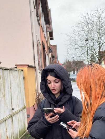 Two females are observed outdoors on a street in Meistratzheim, France, under an overcast sky. The subject positioned centrally on the left has dark hair and wears a black hooded puffer jacket with the hood raised. She holds a smartphone in her left hand and operates its touchscreen with her right index finger; her mouth is slightly open. A backpack strap is visible on her left shoulder. The second subject, partially visible on the right, has bright orange hair and also wears a black puffer jacket. She holds a smartphone with both hands, actively manipulating its screen. A distinct white earring or hair accessory is visible near her right ear. Both individuals are engaged with their respective mobile devices.
The immediate background to the left features a light peach-colored building adjacent to a large, weathered, light-colored double gate. Beyond this, a row of multi-story residential buildings with varying facade colors (peach, brown) and red tiled roofs extends along the street, displaying windows with closed shutters. To the right background, a bare deciduous tree stands, with a black street lamp located behind it. A paved road is visible further back, upon which a light-colored vehicle is present. Additional residential structures, characteristic of European architecture, are discernible in the distance. The scene suggests a cool weather daytime environment.
