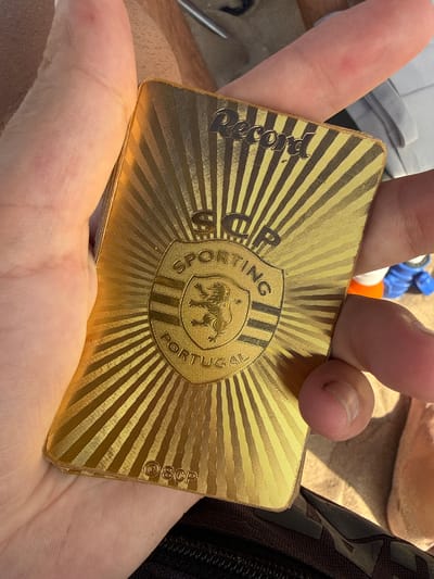 A person's hand is holding a gold-colored card with embossed text and graphics. The card features a sunburst pattern and a crest with a lion. The text "Record," "SCP," "SPORTING," and "PORTUGAL" are visible on the card. A small embossed "SCP" is also present at the bottom. The background is blurred and suggests an outdoor setting. Based on the presence of "Portugal" on the card and its visual characteristics, it is plausible that this item is related to Sporting Clube de Portugal, a Lisbon-based sports club. Without further information or context, it is impossible to definitively determine the specific location within Costa da Caparica, Portugal.