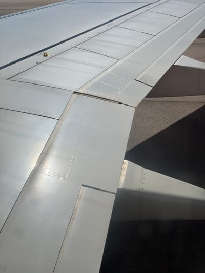 This is an aerial photograph of an airplane wing. The wing is light gray and appears to be made of metal panels joined together with rivets. The wing is positioned above a dark gray surface that is likely tarmac or pavement, as suggested by its textured appearance. A sharp shadow is cast across the tarmac by the wing, indicating sunlight. The photograph is taken from a high angle, looking down at the wing and the ground below. The location in Moreira, Portugal is not discernible from the image itself.