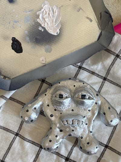 A silver, lumpy creature with black and white polka dots sits on a white and black checkered cloth. The creature has wide-set eyes with dark pupils and visible teeth. To the upper left of the creature is a metallic grey tray with various splotches of paint, including black, white, and light blue. The tray has a grey border.