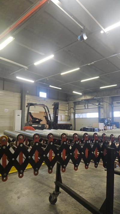 In the city of Tsaratsovo, Bulgaria, an interior shot captures a warehouse or industrial setting. The scene is dominated by a grey concrete ceiling with numerous fluorescent lights illuminating the space. A large, orange-framed overhead door track is visible near the top of the image, suggesting a large bay door. Hanging from the ceiling is a blue and black industrial light fixture. In the mid-ground, an orange and black Toyota forklift is parked. In the foreground, a metal conveyor belt system, designed in an expandable, accordion-like manner with silver rollers and black and orange supports, rests on a black wheeled cart. The floor is concrete. The overall environment is consistent with a storage or manufacturing facility. There are no people visible in the image.