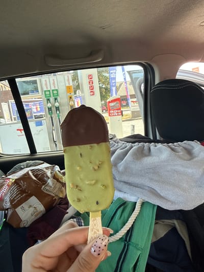 A person's hand with black polka dot nail art holds a chocolate-covered ice cream bar in the passenger seat of a vehicle. The ice cream bar has a dark chocolate top and a light green, pistachio-flavored bottom with visible pistachio pieces. In the background, through the car window, a Total gas station with multiple pumps and signage is visible. The gas station is located on Rue de la Carme in Mont-Saint-Guibert, Belgium. The car interior is cluttered with various items, including a bag of chips, a grey towel, and green and blue clothing. The sky is overcast.