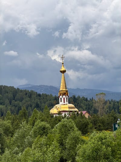 The image depicts a golden-domed church nestled within a lush, green forest. The church's central dome is ornate and topped with a cross. It appears to be a bright, overcast day, with dramatic clouds filling the sky. The foreground is dominated by the dense canopy of trees, with the church peeking out from behind them. In the background, a range of distant, hazy mountains can be seen. The church is located in the village of Chugunash, Russia.