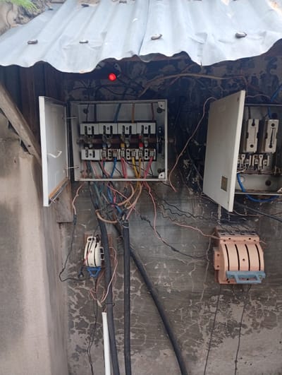 The image shows an outdoor electrical panel installation on a textured concrete wall, likely in Zanzi, Nigeria. A corrugated metal roof is visible at the top, providing partial cover. The main electrical box, open on the left, contains a multi-breaker fuse block with several red and yellow wires connected. A small red indicator light is illuminated above this box. To the right, another electrical enclosure, also open, displays two fuse holders. Below these enclosures, two large, black insulated cables run vertically. A light-colored, rotary-style fuse switch is mounted on the wall between the main enclosures and the cables. Another smaller, white and blue fuse switch is visible further to the left and lower down. The concrete wall exhibits cracks and peeling paint. The scene depicts a functional, albeit somewhat disorganized, electrical setup exposed to the elements.
