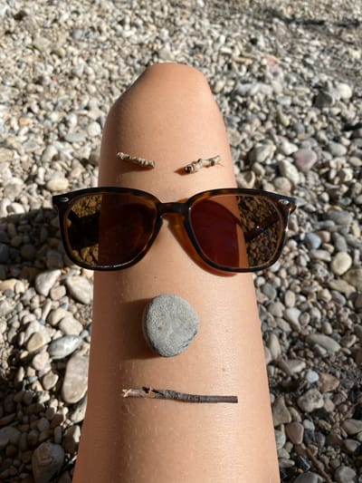 The image shows a person's leg, possibly on a beach or a rocky area, with small natural objects arranged to resemble a face. The leg is tan and has light hair. Sunglasses with dark brown lenses are placed over the middle of the leg. Two small, dry twigs are positioned above the sunglasses to resemble eyebrows. A round, grey pebble is placed below the sunglasses, acting as a nose. A thin, brown twig is positioned horizontally below the pebble, resembling a mouth. The background consists of a dense collection of small, rounded stones of various grey and brown shades. The overall impression is of a playful, anthropomorphic arrangement on a person's limb against a natural, pebbly backdrop. The location in Cortaillod, Switzerland, is not directly discernible from the image content itself.