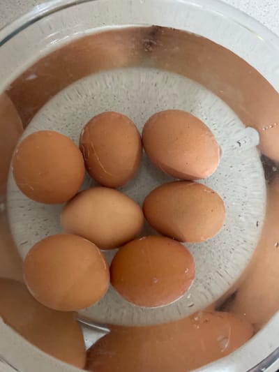 This image shows several brown eggs submerged in water within a clear glass bowl. The eggs are arranged in a circular pattern within the bowl, with some eggs overlapping. The bowl itself has a textured, light gray bottom. The lighting in the image suggests it was taken indoors, possibly in a kitchen. There are some small water droplets visible on the surface of the water and on the eggs. The overall scene is simple and focuses on the eggs in the water. No people are visible in the image. There is no visible text.