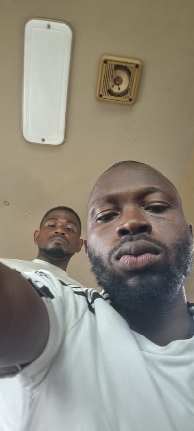 The image, taken in Ilishan, Nigeria, features two men captured from a low-angle perspective, looking down towards the camera. The primary subject in the foreground is a man with dark skin, a full black beard, and short dark hair. He has a neutral expression, with his lips slightly parted, and his eyes are directed at the lens. He is wearing a white shirt, visible on the right side, which appears to have some dark striping or patterns on the sleeve area.
Behind him, positioned towards the lower right, a second man is partially visible. He also has dark skin and a beard, and his gaze is directed downwards at the camera with a neutral expression. He is wearing a white shirt.
The background consists of a plain, light-colored surface, likely a ceiling or an upper wall. Mounted on this surface are two distinct objects. In the upper left corner, there is a square, light-beige fixture with a circular, gridded opening in its center, containing a small, three-pronged element. This object resembles a small speaker or a ventilation unit. Below and to the left of this, a longer, rectangular white fixture is visible, consistent with a light cover or another type of ceiling vent.
The photograph appears to be an indoor shot, possibly a selfie or a photo taken from a very low vantage point. No visible text is present in the image, and the time of day is not discernible.