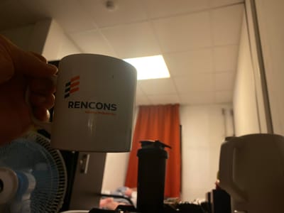 A hand holds a white mug with the "RENCONS Heavy Industries" logo. In the background, a bright overhead light illuminates a tiled ceiling. To the left, a blue oscillating fan is visible. A red curtain hangs behind a black shaker bottle. To the right, the top of a white electric kettle is present. The scene appears to be an indoor setting, possibly a living quarters or common area within the Tecnimont camp in Russia, characterized by functional furniture and diffused lighting.