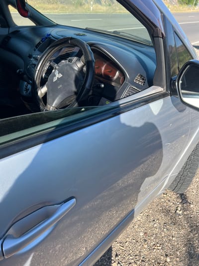 The image shows the interior and exterior of a light gray vehicle parked on the side of a road. The interior includes a dashboard, steering wheel with a Mitsubishi logo, and part of the driver's side door. The exterior shows the driver's side door, door handle, and a portion of the front tire and wheel. The vehicle is parked on a gravel shoulder next to a paved road, with dry vegetation and hills visible in the background. The scene appears to be in a rural or semi-rural area, consistent with the location of Malishka, Armenia. No people are visible in the image. The lighting suggests it is daytime.