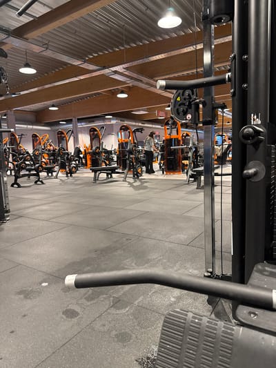 The image depicts the interior of a fitness gym, specifically identified as a Basic-Fit facility located at Rue des Chasseurs Ardennais 2, 6790 Aubange, Belgium. The perspective is from a low angle, partially obscured by a large black fitness machine in the right foreground.
The gym floor is covered with dark grey rubber mats, exhibiting scattered liquid stains or residue, particularly noticeable in the foreground. The ceiling features exposed industrial elements including corrugated metal, light-colored wooden beams, and visible ventilation ducts and electrical conduits. Numerous pendant light fixtures illuminate the space.
The gym contains multiple strength training machines characterized by prominent orange frames and black components, typical of Basic-Fit branding. "Matrix" branding is visible on some equipment. In the right foreground, a black metal frame and a horizontal black bar with a silver end cap are visible, alongside a ridged black footplate, consistent with a cable or weight machine.
Approximately five to seven individuals are present in the mid-ground and background. In the central mid-ground, a female wearing a light-colored long-sleeve top and dark pants stands near an orange and black weight machine, appearing to interact with it. Other individuals are distributed across the gym floor, some seated on or standing near various machines, while others appear to be moving between stations. No direct interactions between individuals are clearly discernible.