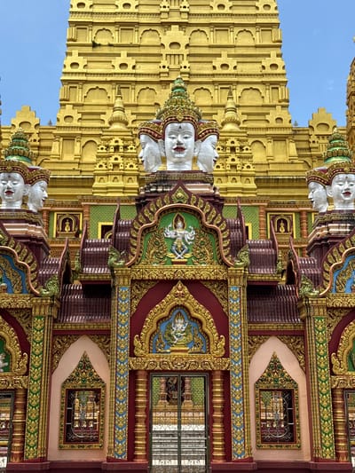 A low-angle, eye-level shot captures the front of a vibrant golden temple in Na Nuea, Thailand. The temple's elaborate facade is adorned with intricate carvings and colorful decorations in shades of red, green, gold, and blue. Dominating the upper portion of the image is a towering, multi-tiered golden structure. In the foreground, four prominent white sculptures of human heads, each with four faces, are mounted on ornate structures. These heads are topped with decorative headdresses, and their serene expressions gaze outwards. Between these sculptures, decorative arched entrances and windows are visible, filled with patterned metal grates. The sky above is a clear, pale blue.