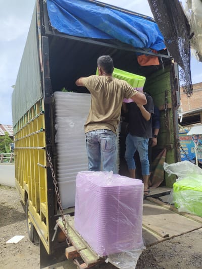 A man in a tan shirt and blue jeans is loading a stack of purple plastic containers into the back of a yellow truck. He is facing away from the camera and appears to be placing the containers on top of other stacked items. To his left, another man in a black shirt and blue jeans is also inside the truck, assisting with the loading. The truck is open at the back, revealing a large quantity of white plastic containers stacked inside. There are also green plastic containers visible. The truck is parked on a dirt road next to a building with a sign in a foreign language, possibly a business. The background shows trees and a cloudy sky. The overall scene suggests a commercial delivery or relocation of goods in Keudeblangme, Indonesia.