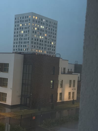 The image shows a cluster of modern buildings in Korobovo, Russia. In the background, a tall, white, multi-story apartment building dominates the scene. Its facade is punctuated by numerous windows, many of which are illuminated, suggesting occupancy. In the foreground, a lower, white building with a section of dark brick cladding is visible. This building also features windows, some of which are lit. The area between these buildings appears to be a courtyard or open space. A dark metal fence runs across the lower part of the image, separating the buildings from an area with some vegetation. The sky is overcast and a dull, muted blue, indicating it is likely dusk or a heavily clouded day. There are no people visible in the image. The overall impression is one of an urban residential area.