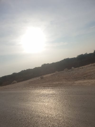 The image captures an outdoor landscape scene, likely in Killi, Syria, under the intense glare of the sun. The perspective is oriented with a very bright sun dominating the left side of the frame, appearing low in the sky, which suggests either early morning or late afternoon. The sky, occupying the upper and left portions, is a hazy, light blue-grey, with some indistinct cloud formations partially visible through the strong sunlight.
A dark, irregular line representing distant hills or a treeline stretches diagonally across the middle of the frame, forming the horizon and separating the bright sky from the ground below. The right and lower portions of the image reveal a wide, textured surface, which appears to be an unpaved road or a broad path. This granular surface exhibits significant specular reflection, indicating either a wet ground surface or the powerful glint of the low sun reflecting off its texture.
No people, animals, or distinct man-made objects beyond the possible road are visible. The scene portrays a serene, somewhat desolate landscape dominated by natural elements and strong sunlight. There is no visible text within the image.