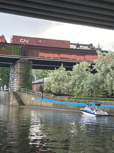 The image shows a view from a body of water, likely the Sainte-Anne River or Lake Saint-Louis, looking towards a railway bridge in Sainte-Anne-de-Bellevue, Canada. A CN train car, maroon with white lettering, is visible on the bridge. The bridge itself is supported by large stone pillars covered in ivy. Below the bridge, a concrete embankment lines the water, featuring a set of stairs leading up from the water's edge. A small building with a "VHF 68" sign is situated behind the embankment. To the right, lush green trees with light-colored foliage are abundant. A white and blue motorboat with a navy blue canopy is docked near the embankment. The sky is overcast, and the water reflects the sky and surrounding elements.