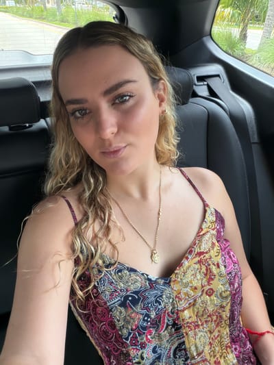 A young woman is reclined in a black car seat, presumably in the back of the car, as indicated by the positioning and surrounding interior features. She is wearing a patterned top with thin straps, a gold necklace, and a red bracelet on her arm. Her hair is blonde and curly, and she is looking toward the camera with a neutral expression. Outside the car window, green foliage is visible, suggesting an outdoor, potentially tropical or subtropical environment. Based on the prompt, the location is Key Biscayne, United States of America.