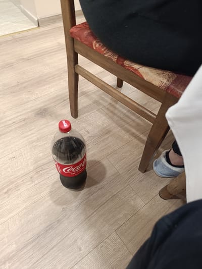 An indoor scene shows a partly consumed 1.5 liter bottle of Coca-Cola Original Taste standing on a light wood-grain floor next to a wooden chair with a patterned cushion, possibly located in Cherkovitsa, Bulgaria. A person, wearing dark clothing, sits on the chair. Another person, also in dark clothing, is visible in the lower right. One foot, wearing a blue slipper, is visible near the chair leg.