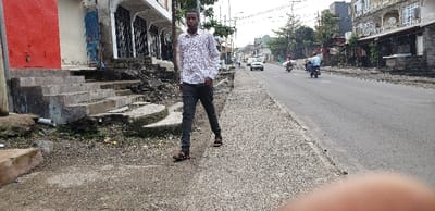A young man in a white and black patterned shirt and blue jeans walks on the sidewalk. He is wearing sandals. He is walking away from the camera towards a paved road. The sidewalk is made of loose gravel and there are a set of concrete steps leading up to a building in the background. The road is paved and there are multiple vehicles and people in the distance. There are buildings and trees on both sides of the road. The photo was taken in Keshero, Democratic Republic of the Congo. The coordinates are -1.664 latitude and 29.181 longitude.