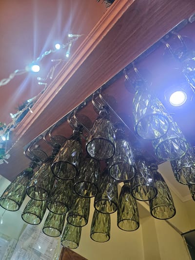 An upward-angle, eye-level shot captures a rack of green, faceted wine glasses hanging upside down from a wooden shelf. String lights with white bulbs are draped above the glasses, creating a festive atmosphere. A bright light source, possibly a bulb, is visible in the upper right corner, casting a strong blue-white glare. The background is blurred, but hints of a wall and possibly other shelving or decorations can be discerned. There are no people visible in the image. The setting appears to be indoors, possibly a bar or dining area. The specific location in Skadovsk, Ukraine, cannot be determined from the visual information provided.