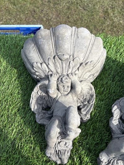 The image shows a stone angel statue lying on artificial green grass. The statue has a stylized shell or fan behind its back and another angel statue is partially visible in the upper right. A blue plastic container is visible to the left. The location is Chirk, United Kingdom.