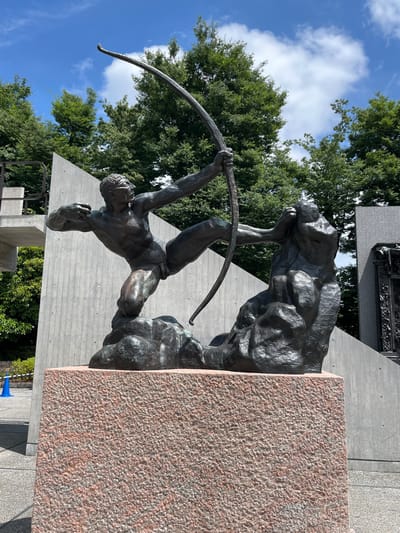 The image depicts "The Archer" (Héraklès archer), a bronze sculpture by Émile-Antoine Bourdelle, situated in Ueno Park, Taito, Tokyo, Japan, specifically outside the National Museum of Western Art.

The sculpture portrays a muscular male figure, identified as Hercules, in a dynamic and highly tensed pose, kneeling on a craggy bronze base while drawing a large, unstrung bow. His body is twisted, with his left leg bent forward and his right leg extended back. His arms are positioned to pull the bow, his right hand gripping the string area and his left hand holding the bow itself. His facial expression appears strained, and his eyes are fixed towards an unseen target. To the right of the archer, another, more abstract rocky bronze mass rises from the same base. The sculpture rests on a substantial rectangular pedestal of coarse, reddish-pink granite.

In the background, on the left, a tall, angled concrete wall with a raw, unfinished texture is visible. Further to the left, a light grey concrete structure with railings extends, suggesting an elevated walkway or architectural element. A blue traffic cone is partially visible on the concrete ground in the lower-left corner. On the right side of the background, another, slightly darker and textured concrete wall is present. Partially visible to the far right is a large, intricate dark bronze sculpture or architectural facade, characteristic of "The Gates of Hell" by Auguste Rodin, also located at the museum. Lush green foliage from various trees frames the sculpture against a bright blue sky with scattered white cumulus clouds, indicating an outdoor setting on a clear day. The ground immediately surrounding the granite pedestal is paved with light grey concrete.