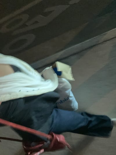 The image shows an array of items haphazardly arranged. A white garment, possibly a dress, drapes downward with visible detailing. Next to it, a cardboard box sits beside a white plastic bag printed with "Columbia." Dark blue pants are also present, alongside a red metal object, possibly a decorative element or tool. The backdrop features a dark surface with large, blurred letters.