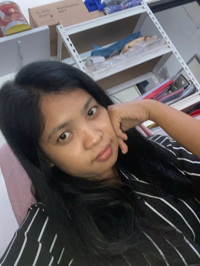 A person with dark hair and fair skin is pictured indoors. The individual is wearing a black and white striped shirt and resting their head on their hand. The background includes shelves containing various items, including clothes and boxes. The specific location in Negara, Indonesia is not discernible from the image.