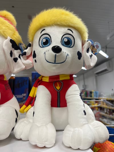 The image shows two stuffed dogs, likely from the Paw Patrol franchise, displayed on a shelf. The dog in the foreground is white with black spots on its ears and legs, yellow fur on its head, a red vest with a yellow and red striped scarf. The dog has blue eyes and a black nose. A tag with Paw Patrol characters is visible on its ear. The background is blurred but shows shelves stocked with various items, including boxes with blue labels. An air conditioner is visible on the wall in the background. The lighting suggests an indoor retail environment. The image does not provide any information about the city of Ivanivka, Ukraine.