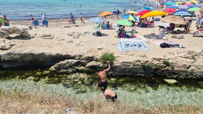 The image depicts a sunny day at a crowded beach in Porto Cesareo, Italy. In the foreground, two young males are wading in shallow, clear water near a rocky, vegetated embankment. One of them is reaching up towards a bushy plant on the rocks. The middle ground features a sandy beach with numerous beach umbrellas and sunbathers. Several people are seen walking along the shoreline carrying beach chairs and bags. In the background, the turquoise sea is dotted with swimmers and boats, with the distant coastline visible. The overall scene conveys a lively and typical summer beach atmosphere.