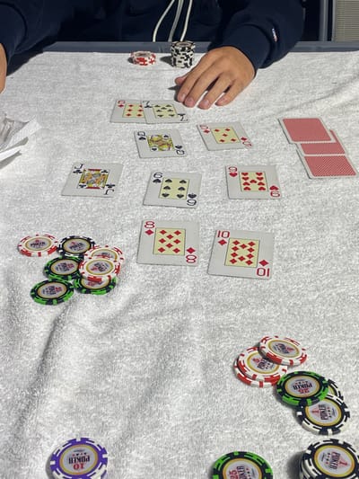 The image captures an ongoing poker game played on a white terry cloth towel covering a grey table surface in Yavne, Israel. A person's forearms and hands, wearing a dark blue long-sleeved top with a white zipper and a subtle emblem on the right sleeve, are visible at the top, with the right hand resting on the towel near a stack of black and white poker chips and a single red and white chip. Various poker chips in red/white (value 1), green/black (value 25), and purple/white (value 10) are distributed across the table in multiple clusters. Worn playing cards are face-up: a hand of 6 of Diamonds and 7 of Clubs is at the top center; a hand of Jack of Clubs and 6 of Spades is mid-left; a hand of Queen of Spades and 5 of Diamonds is mid-right; and another hand featuring 9 of Hearts and 10 of Hearts is further right. Two communal cards, 8 of Diamonds and 10 of Diamonds, are centered in the lower portion of the image. A partial deck of red-backed playing cards rests at the top right, and a crumpled white object, possibly a disposable mask or napkin, is present on the far left.