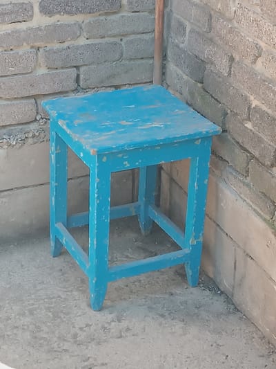 A weathered, bright blue wooden stool stands on a concrete surface next to a grey brick wall. The stool has a square seat and four legs connected by stretchers. The paint on the stool is chipped and peeling, revealing the wood underneath. The brick wall behind the stool is constructed of rough, rectangular grey blocks, some of which appear to be crumbling or covered in a fine layer of dust or dirt. A vertical, thin brown pole or pipe is visible to the right of the stool, partially obscured by the stool and the wall. The scene is outdoors, with the lighting suggesting it is daytime. The environment appears to be a utilitarian or possibly abandoned area. The location is identified as Yasnohirka, Ukraine. There are no people or animals present in the image.