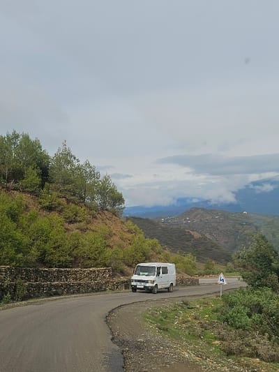 The image displays a winding paved road through a mountainous landscape in the vicinity of Moqrisset, Morocco, under an overcast sky. A white panel van, resembling a Mercedes-Benz T1 model, is centrally positioned on the asphalt road, traveling away from the camera. The van features dark-tinted windows in its cargo area. To the left of the road, a multi-tiered dry-stone retaining wall supports a steep, tree-covered hillside with numerous green evergreen trees and shrubs. To the right of the road, a blue and white warning sign indicating a series of curves (depicted as an 'S' shape) stands amidst sparse green vegetation. In the background, undulating hills and mountains stretch into the distance, with some peaks partially obscured by low-lying clouds or fog. No individuals are visible in the scene.