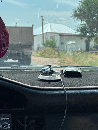This media file depicts a close-up view of a car's dashboard, seen from inside the vehicle. The dashboard is covered with a dark, textured material. On the dashboard, there are several objects, including an air freshener with a spinning propeller, and a small black electronic device. A wire extends from the air freshener. Through the windshield, a blurred outdoor scene is visible. This includes trees, buildings, and what appears to be a truck or large vehicle. The lighting suggests it is daytime. In the lower right corner of the image, a small black object with white text is visible; the text reads "areon QUALITY PERFUME" and includes some circular imagery. The location context provided is Сарымолдаев ауылдық округі, Kazakhstan.