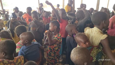 A group of African children and adults are gathered in a large, open-air structure, likely a community center or shelter, in Bunia, Democratic Republic of the Congo. The lighting suggests it is daytime, possibly indoors with natural light filtering in. Many of the children appear to be young, with some infants held by older children or adults. Their clothing is varied, ranging from simple t-shirts and dresses to more traditional patterned fabrics. Some individuals are looking directly at the camera, while others are engaged in conversation or looking elsewhere. The environment appears somewhat rustic, with visible wooden support beams and a dirt floor. The overall impression is one of a community gathering, possibly related to an event, aid distribution, or a social occasion. The bottom right corner of the image contains a watermark with the text "Joseph Kisuki".