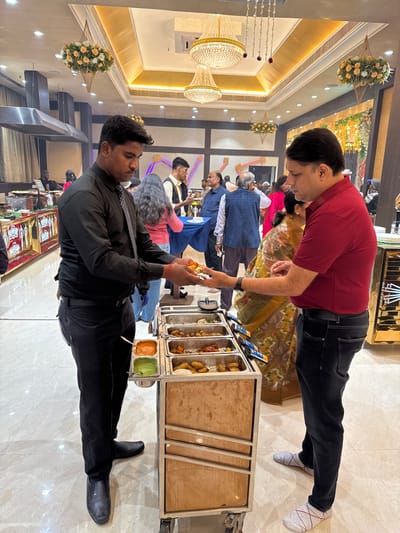 Inside a well-lit banquet hall, a man in a maroon shirt and dark jeans receives a snack from a server in a black uniform. A buffet cart with various food containers and sauces stands between them. Other guests are milling about in the background. The hall features ornate chandeliers, floral decorations, and a decorated ceiling. The floor appears to be polished tile.